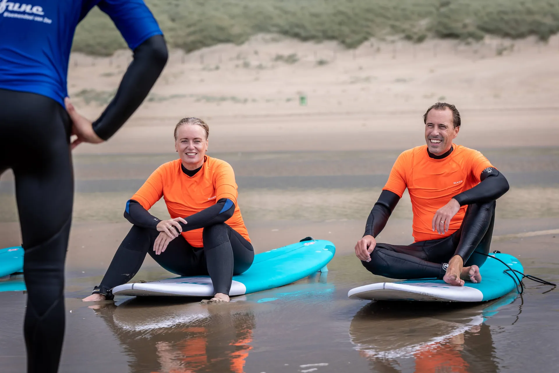 Twee partners zitten lachend op surfplanken aan de waterkant