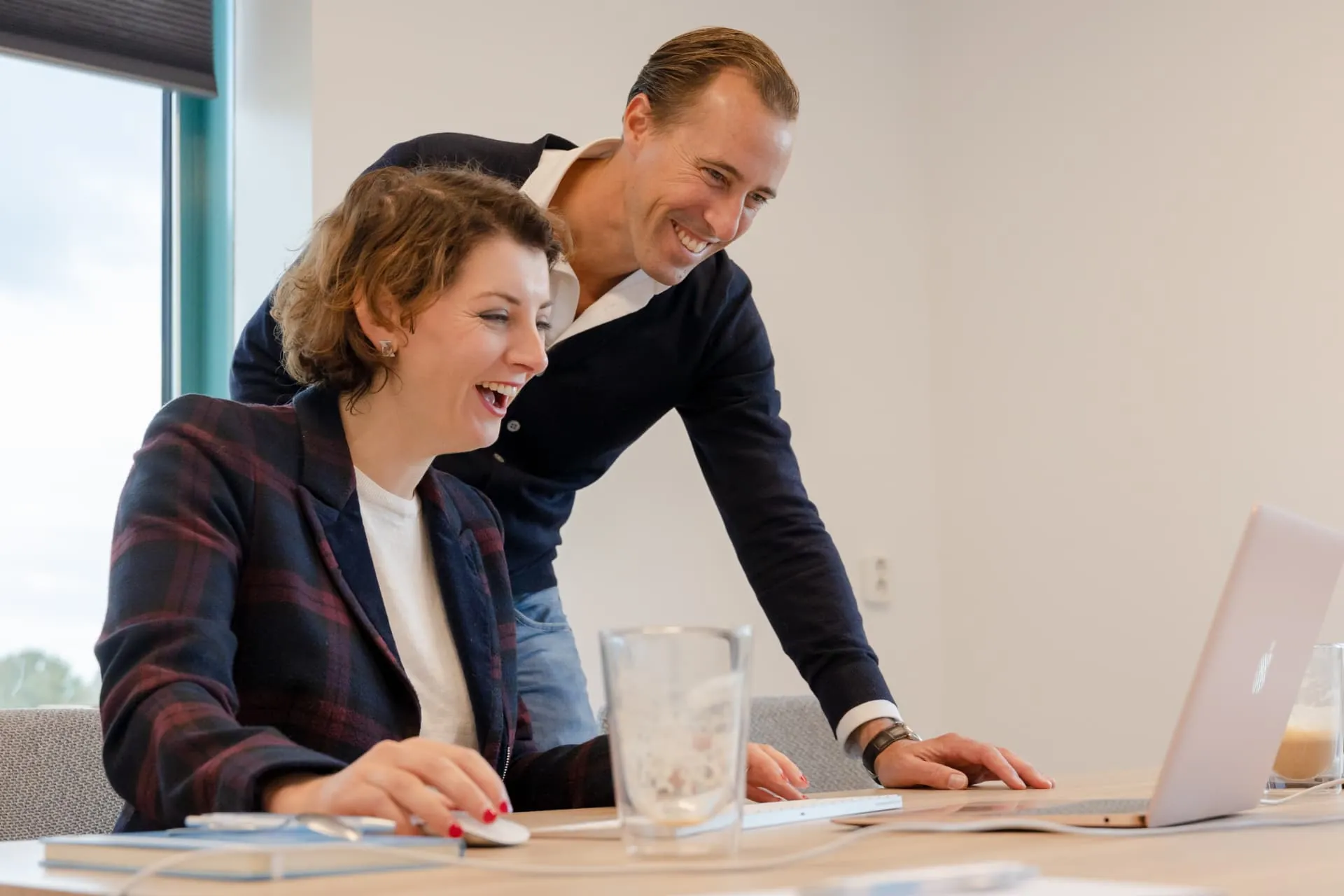 Twee collega's kijken lachend mee op een laptopscherm in een licht kantoor